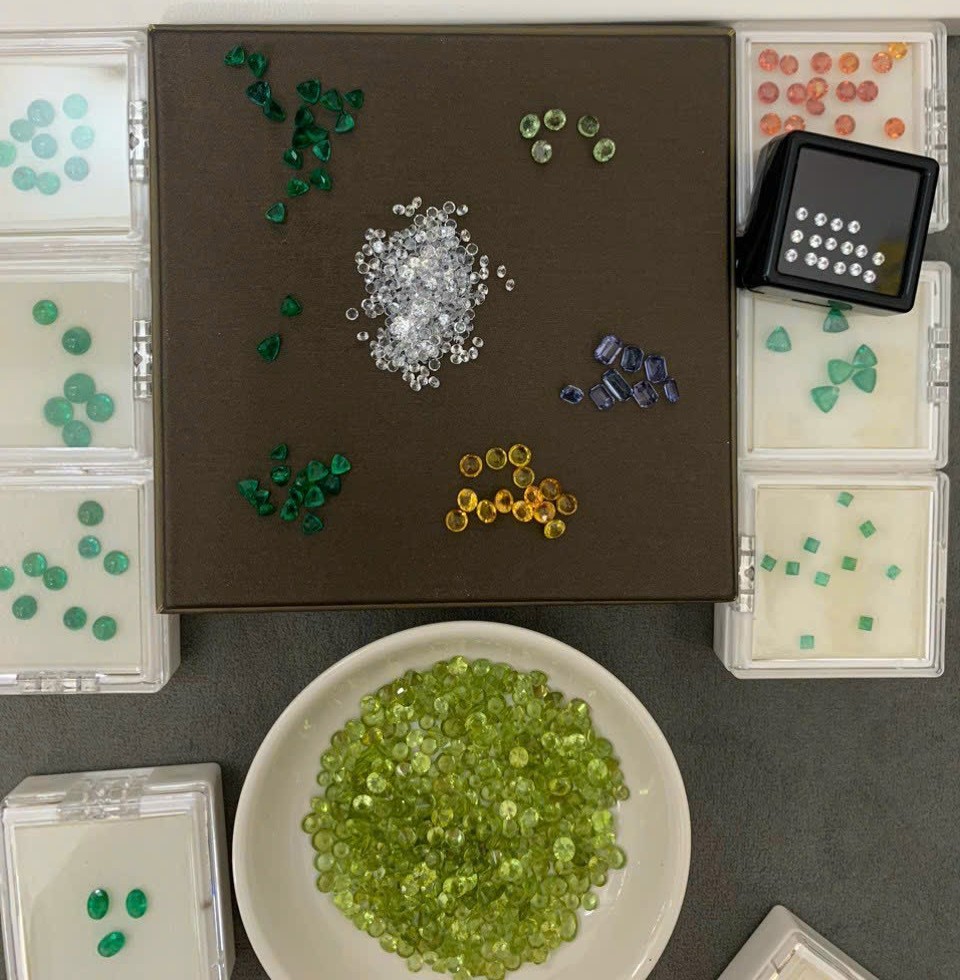 A dealer sorting table with a dish of small round peridot beside parcel boxes of emerald, sapphire and other coloured stones
